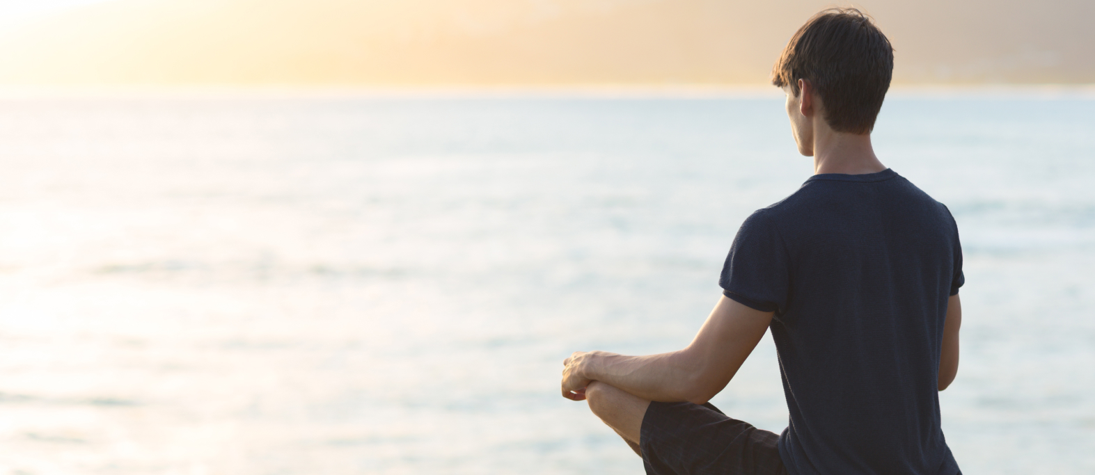 A person sits cross-legged on a rocky edge overlooking the ocean at sunset, meditating and embracing mindfulness. This image represents relaxation and self-care while travelling with mental health conditions, highlighting the importance of peaceful moments during a journey